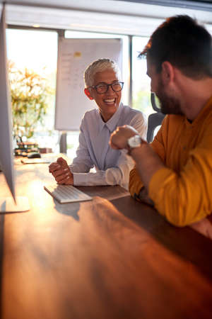 An elderly business woman has a good time with her young male colleague while they working at the desk in pleasant atmosphere in the office. Business, office, jobの写真素材