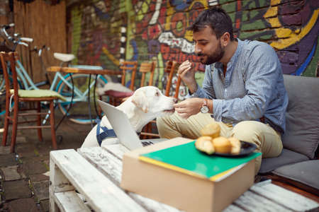 A young male student is feeding his dog while spending free time at the bar's garden on a beautiful day. Leisure, bar, friendship, outdoorの写真素材