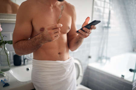 close up of young adult caucasian male torso, holding his cell phone and a tooth brush in the bathroom, half nakedの写真素材