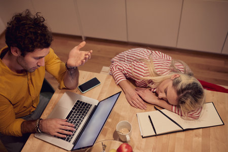 young adult caucasian male chatting online while his girlfriend fell asleep at the kitchen tableの写真素材
