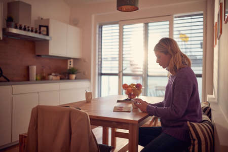 young adult  caucasian blonde female reading content on her cell phone, sitting in sunny kitchenの写真素材