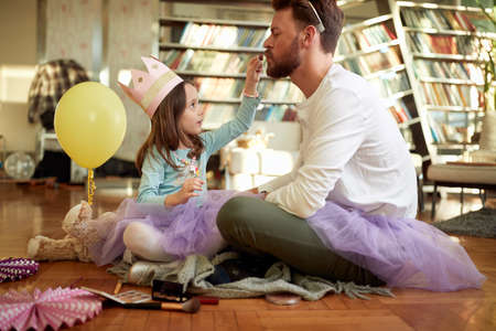 A little daughter is putting the lipstick on the lips of her father while they preparing for a ballet training in a relaxed atmosphere at home. Family, together, homeの写真素材