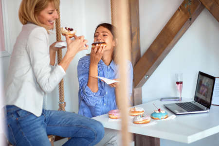 Two female friends in a pastry shop are passionately eating delicious donuts of irresistible...