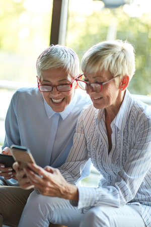 Two elderly business women of similar appearance enjoy a smartphone content at the break in a pleasant atmosphere at workplace. Business, office, jobの写真素材