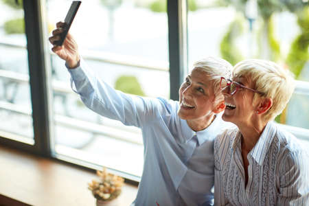 Two older business women of similar appearance have a good time taking selfies during a break in a cheerful atmosphere at workplace. Business, office, jobの写真素材