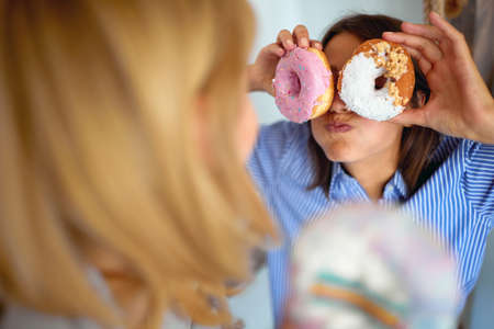 A girl making funny grimaces using delicious donuts while spending time with her friend in a cheerful atmosphere in a pastry shop. Pastry shop, dessert, sweetの写真素材