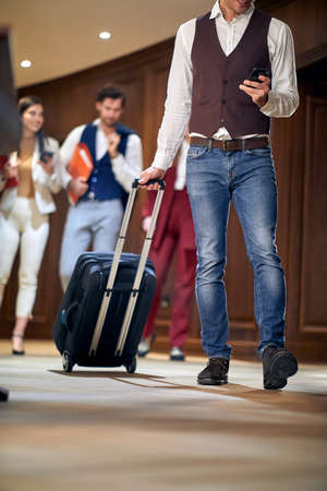 group of young adults, business people, walking through the hallway. Selective focus on a male with cell phone, carrying wheeled suitcase. Cropped imageの写真素材