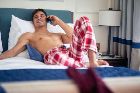 half naked caucasian young male talking on his cell phone, smiling, looking happy, relaxed. Coffee cup on bedside table.の写真素材