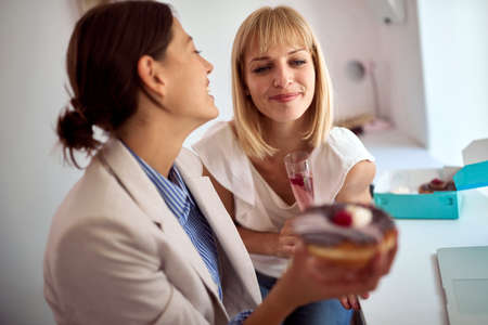 Two happy female coworkers eating donuts together on the break