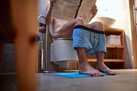 A young man sitting on the toilet in a relaxed atmosphere in the bathroom and reading a newspapers. Toilet, need, homeの写真素材