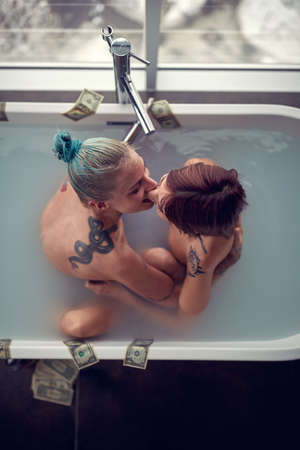 A young sexy tattooed lesbian couple kissing and hugging while having a warm bath in a relaxed atmosphere at the bathroom together. Bathroom, relationship, love, togetherの写真素材