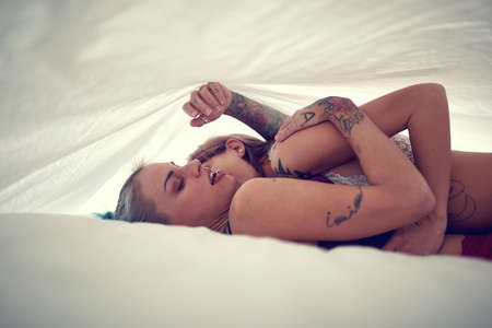 Young sexy tattooed lesbian couple full of passion having a sex in an intimate atmosphere under the white sheet. Love, relationship, sex, lgbtの写真素材