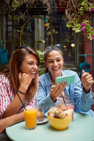 one female friend showing to another something on her cell phone sitting in outdoor cafeの写真素材