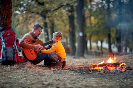 Grandfather and grandson enjoying playing a guitar around campfire in the forest on a beautiful...