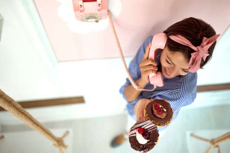 Closeup from above of a young beautiful girl chatting on the phone in a pleasant atmosphere in a retro looking pastry shop while holding chocolate donuts in her hand. Pastry shop, dessert, sweetの写真素材