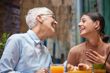 caucasian mother and daughter sitting in outdoor cafe, looking each other, smiling. hand on chest,...