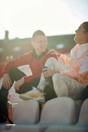 Smiling woman jogger in sportswear sitting on tribune talking with young runner man