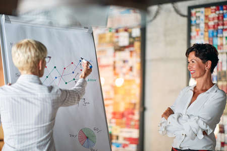 An elderly business woman is enjoying in her female colleague graph explanation in a pleasant atmosphere at workplace. Business, office, jobの写真素材