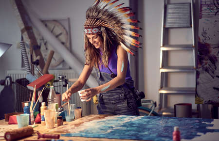 A young female artist with a war bonnet working on a new painting in a pleasant atmosphere in the studio. Art, painting, studioの写真素材