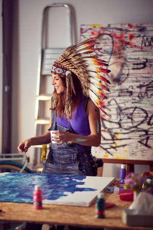 A young female artist with a war bonnet is in a good mood while working on her new painting in a pleasant atmosphere in the studio. Art, painting, studioの写真素材
