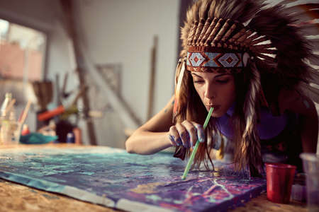 A young female artist with a war bonnet is using a straw technique while working on a new painting in a creative atmosphere in the studio. Art, painting, studioの写真素材
