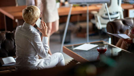 A senior business woman sitting on the sofa and talking on the smartphone while listening to a female colleague presentation in a pleasant atmosphere at workplace. Business, office, jobの写真素材