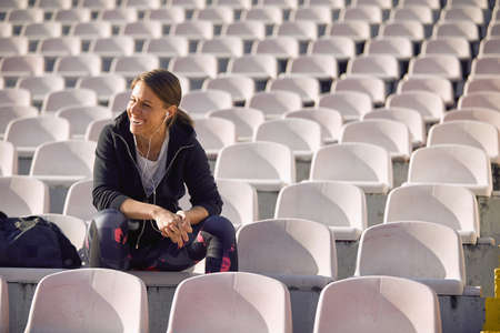 A young woman in a good mood is sitting on the grandstand and relaxing after training on a beautiful day at the stadium by listening the music. Sport, athletics, athletesの写真素材