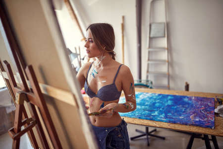 A young sexy and attractive female artist in a bra is standing in front of the easel in a relaxed atmosphere in her studio and painting. Art, painting, studioの写真素材