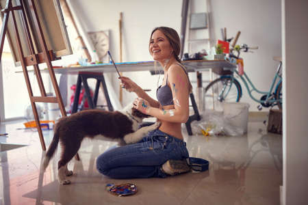 A young sexy and attractive female artist in a bra is in a good mood in a relaxed atmosphere in the studio while playing on the floor with her dog. Art, painting, studioの写真素材