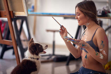 A young sexy and attractive female artist in a bra is playing with her dog while taking a break from painting in a relaxed atmosphere in her studio. Art, painting, studioの写真素材