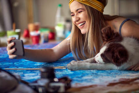 Close-up of a sexy and attractive female artist is standing over the worktable in a relaxed atmosphere in her studio and taking a selfie with her dog. Art, painting, studioの写真素材