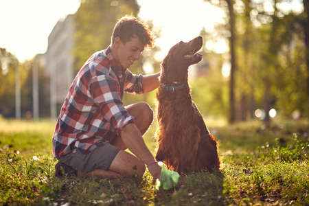 A young guy is kneeling on the grass on a beautiful day in the park and collecting the poop of his dog. Friendship, walk, petsの写真素材