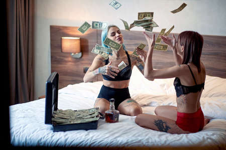 A young sexy tattooed lesbian couple in the lingerie is sitting on the bed in a cheerful atmosphere and enjoy throwing the money in the air. Home, relationship, loveの写真素材
