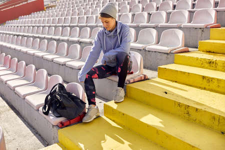 A young girl is sitting on the grandstand and concentrating for a training on a beautiful day at the stadium by listening to a music. Sport, athletics, athletesの写真素材
