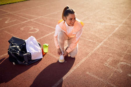 caucasian young adult female kneeling on one knee at the athletic track, tying shoelaces, smiling, looking  away with her headphones in her earsの写真素材