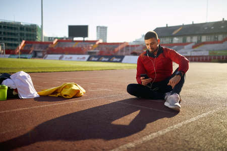 young adult caucasian beardy man sitting on athletic track at stadium in sportswear, looking at his cell phoneの写真素材