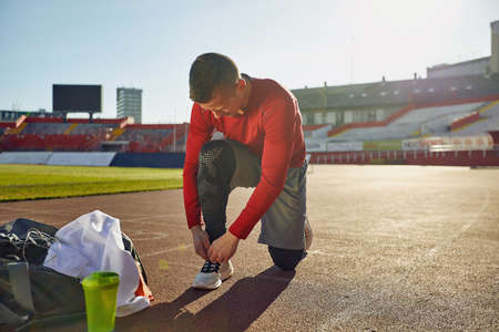 young adult caucasian male kneeling on one knee at the stadium athletic track, tying shoelaces, preparing for trainingの写真素材