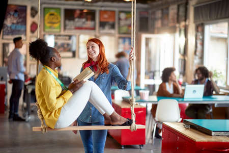 A young woman is chatting with her young female colleague while relaxing on a swing during a break in a relaxed atmosphere in the office. Employees, office, workの写真素材