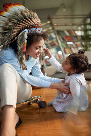A young Mom with an indian headdress spending quality time with her little daughter in a family atmosphere at home. Family, together, love, playtimeの写真素材