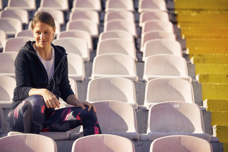 beautiful young adult caucasian female sitting at the stadium  with headphones in her ears, smiling, talking on her cell phoneの写真素材
