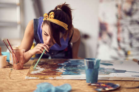 Close-up of a young female artist using a straw while working on a new painting in a creative atmosphere in the studio. Art, painting, studioの写真素材