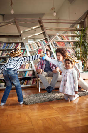 Mom and her son yelling and playing with swords in a cheerful atmosphere at home while Dad and a little daughter enjoy watching. Family, together, love, playtimeの写真素材