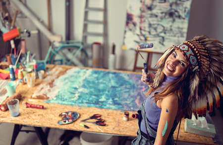 A young female artist with a war bonnet in a pleasant atmosphere in the studio is posing for a photo while working on a new painting. Art, painting, studioの写真素材