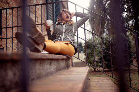 A young handsome woman yawns while sitting on the stairs in front of the house and enjoying a book and a coffee in a relaxed atmosphere. City, outside, restの写真素材