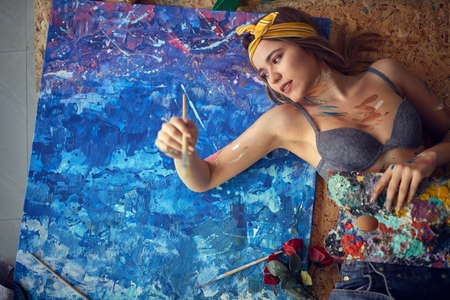 A sexy and attractive female artist is lying on the floor in a relaxed atmosphere in her studio and working on the painting. Art, painting, studioの写真素材