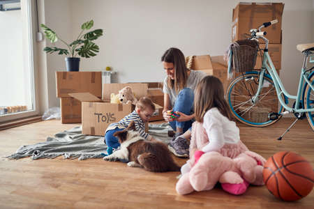 young mom playing with her son, daughter and a dog on the wooden floor in new apartment with unpacked boxes around them.の写真素材