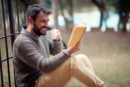 A young handsome man is reading a book and drink a coffee while sitting on the stairs in front of the house in relaxed atmosphere. City, outside, restの写真素材