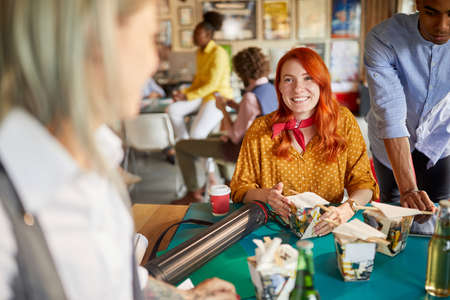 cute caucasian redhead female talking and smiling to a colleagues at work, having lunch break, eating chinese foodの写真素材