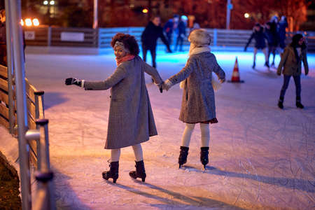 A shot from the back of two female friends while skating at ice-skating rink during Christmas holidays on a beautiful night  in the city. Christmas, New Year, holiday, friendshipの写真素材