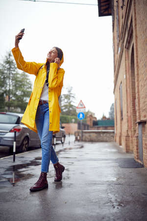A young cheerful woman in a yellow raincoat is listening to the music while walking the city in a good mood on a rainy day. Walk, rain, cityの写真素材